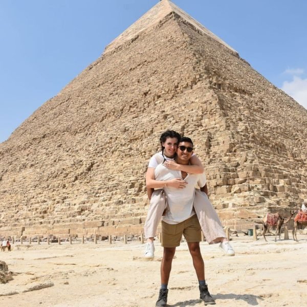 a wonderful shot of a husband carrying his wife on his back in front of the giza pyramids