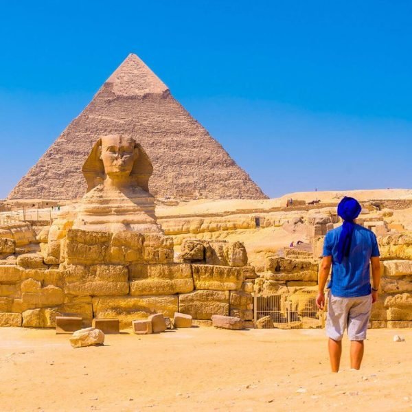 traveler standing before the Great Sphinx with the Pyramids of Giza rising in the background under a clear sky.
