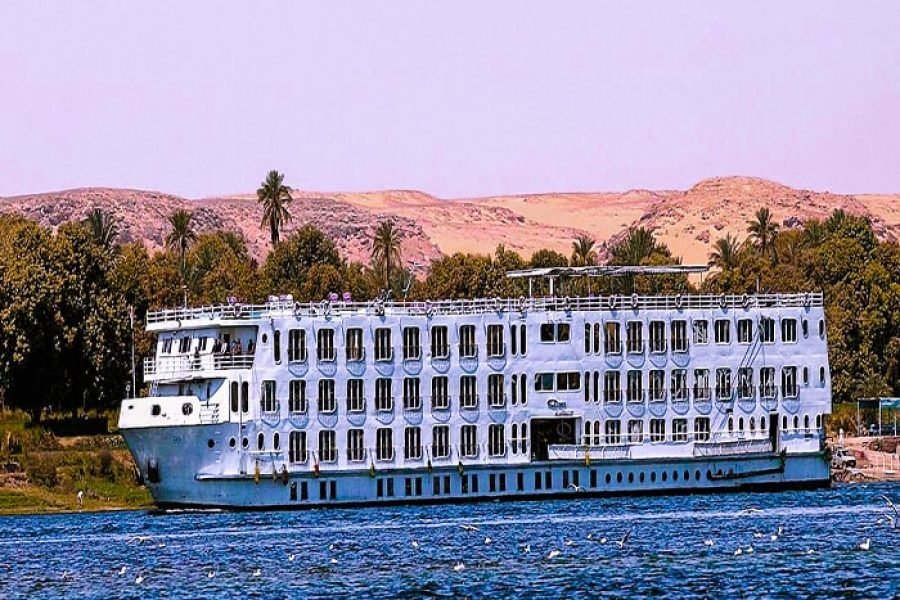 Full view of the Nile Quest cruise ship as it sails past palm trees along the Nile River.