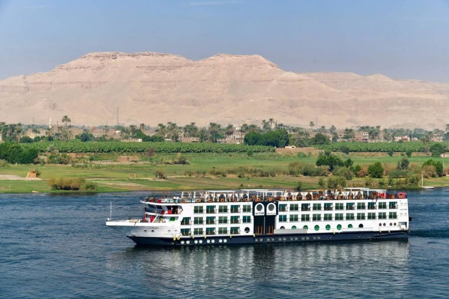 Exterior of the Mayfair Nile cruise ship sailing past the lush riverbanks and arid desert mountains.