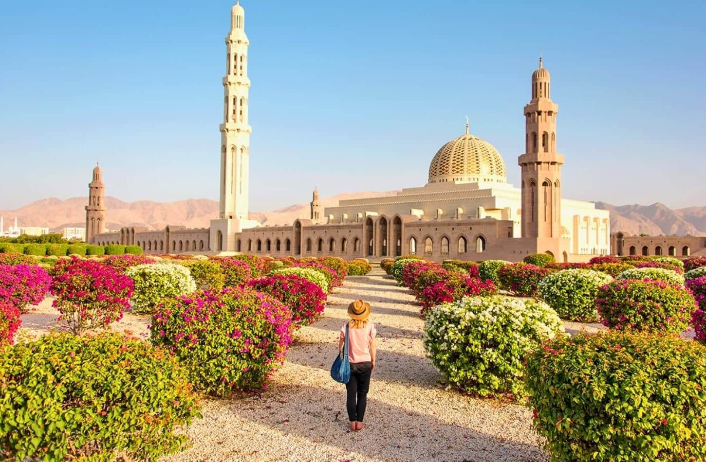 A Woman Tourist walking to a grand mosque, flanked by vibrant flowers, during the best time to visit Oman.