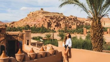 A woman relaxing under the sun with Ait Ben Haddou in the background, showing that it is safe to travel to Morocco and explore its beauty.