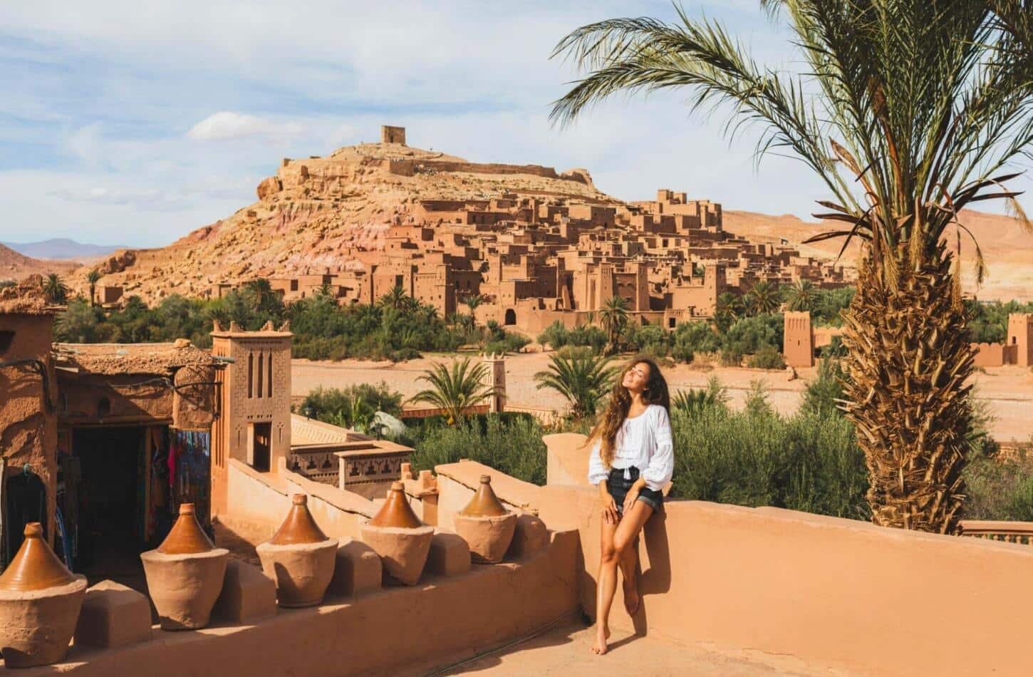 A woman relaxing under the sun with Ait Ben Haddou in the background, showing that it is safe to travel to Morocco and explore its beauty.