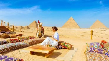 A woman enjoys sitting by the pyramids with a decorated camel in Christmas attire and colorful cushions around her, perfectly capturing the spirit of Egypt Christmas holiday packages.