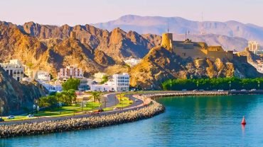 panoramic view of a curved coastal road and blue water next to rugged mountains and an ancient stone fort