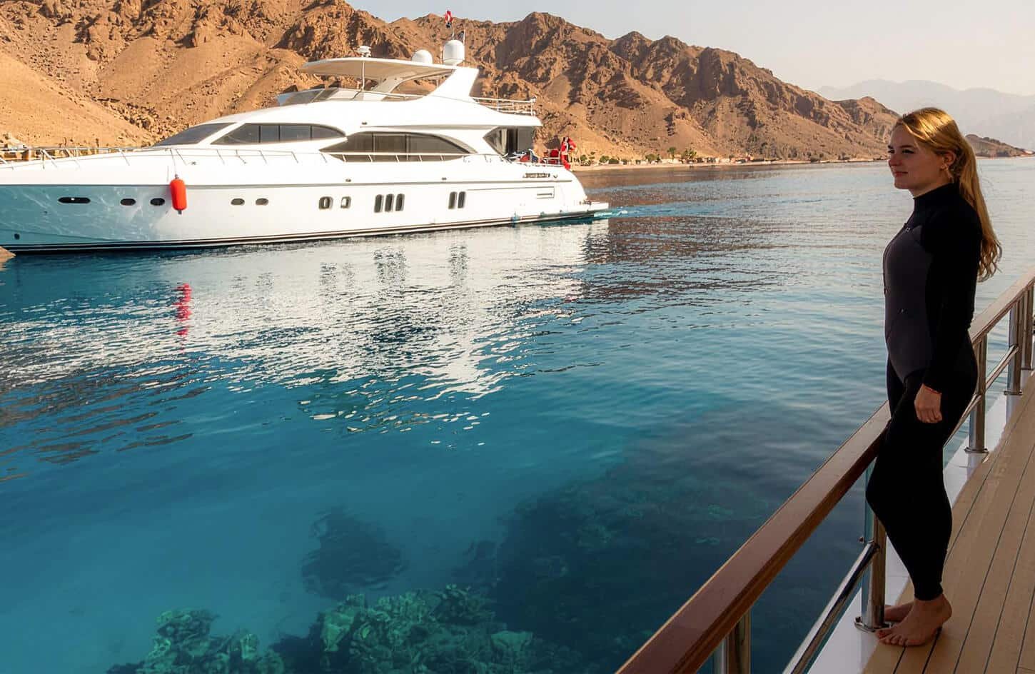 Woman in a wetsuit on a yacht deck, ready to dive into the clear Red Sea water near mountains