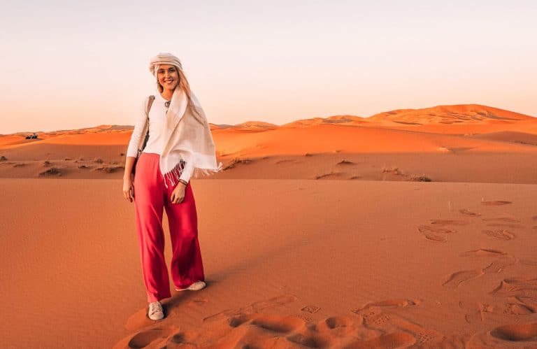Smiling woman in pink pants on Moroccan Sahara sand dunes at sunset.