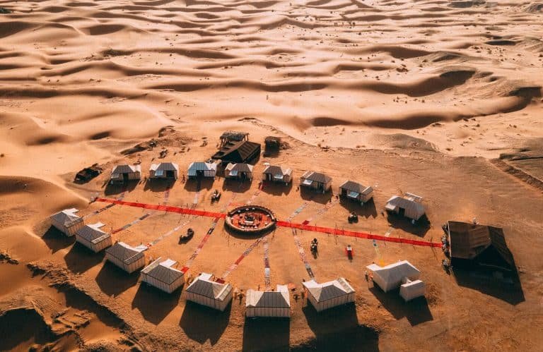 Aerial view of a luxury, circular Moroccan desert camp with tents and red carpets on the Sahara sand dunes.