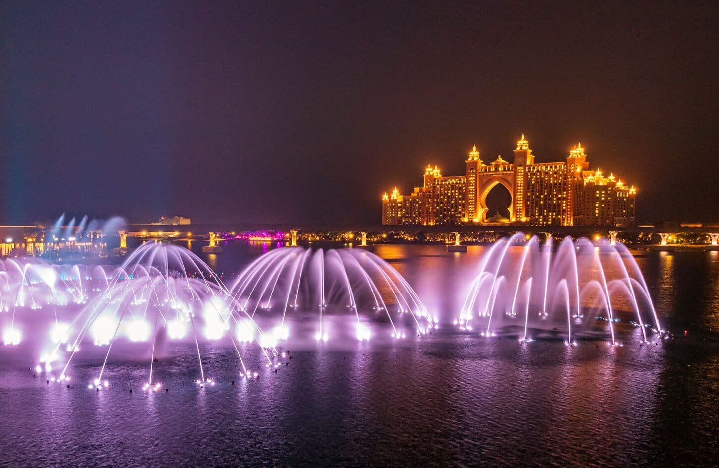 Purple-lit Dubai fountain night show with the brightly illuminated Atlantis, The Palm hotel in the background.