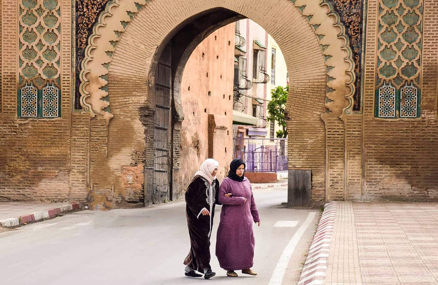 Two women in djellabas walking through the ornate, tiled, arched city gate of Meknes, Morocco.