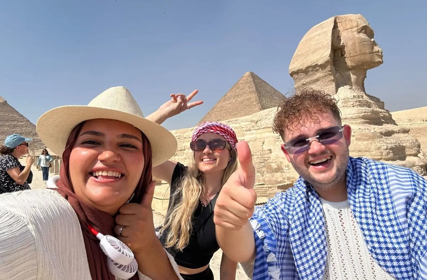 Three smiling people posing for a selfie in front of the Sphinx and ancient Egyptian pyramids in the desert.