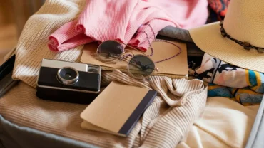 A flat lay of an open suitcase organized for a Packing List for A Nile Cruise, featuring a vintage-style camera, round sunglasses with a purple chain, a straw sun hat, and folded clothing in cream and soft pink.